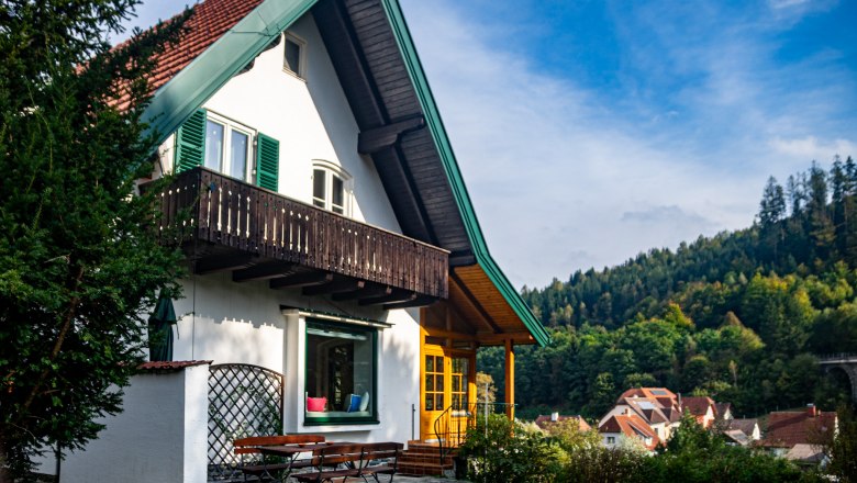 Ein weißes Haus mit rotem Dach und grünem Balkon vor einem bewaldeten Hügel.