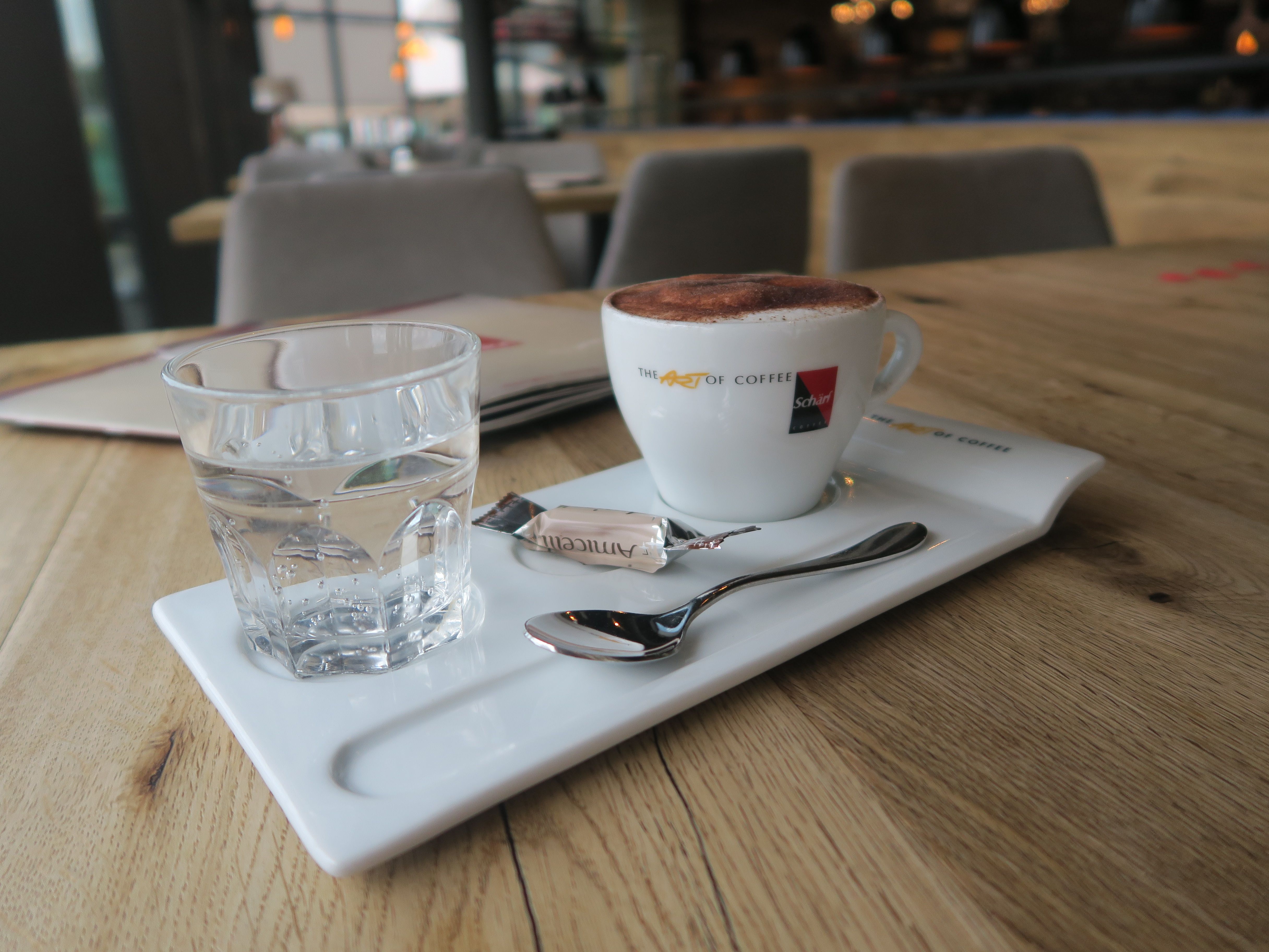 Eine Tasse Kaffee mit Schaum, ein Glas Wasser und ein Löffel auf einem Tablett in einem Café.