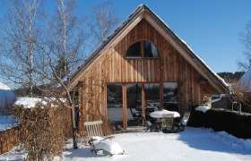 Ein Holzferienhaus im Schnee mit Gartenm&ouml;beln und kahlen B&auml;umen.
