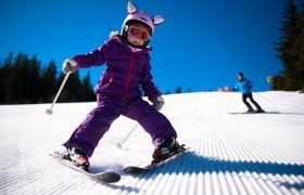 A child in a purple ski suit is skiing down a groomed slope, followed by an adult. The sky is clear and blue.
