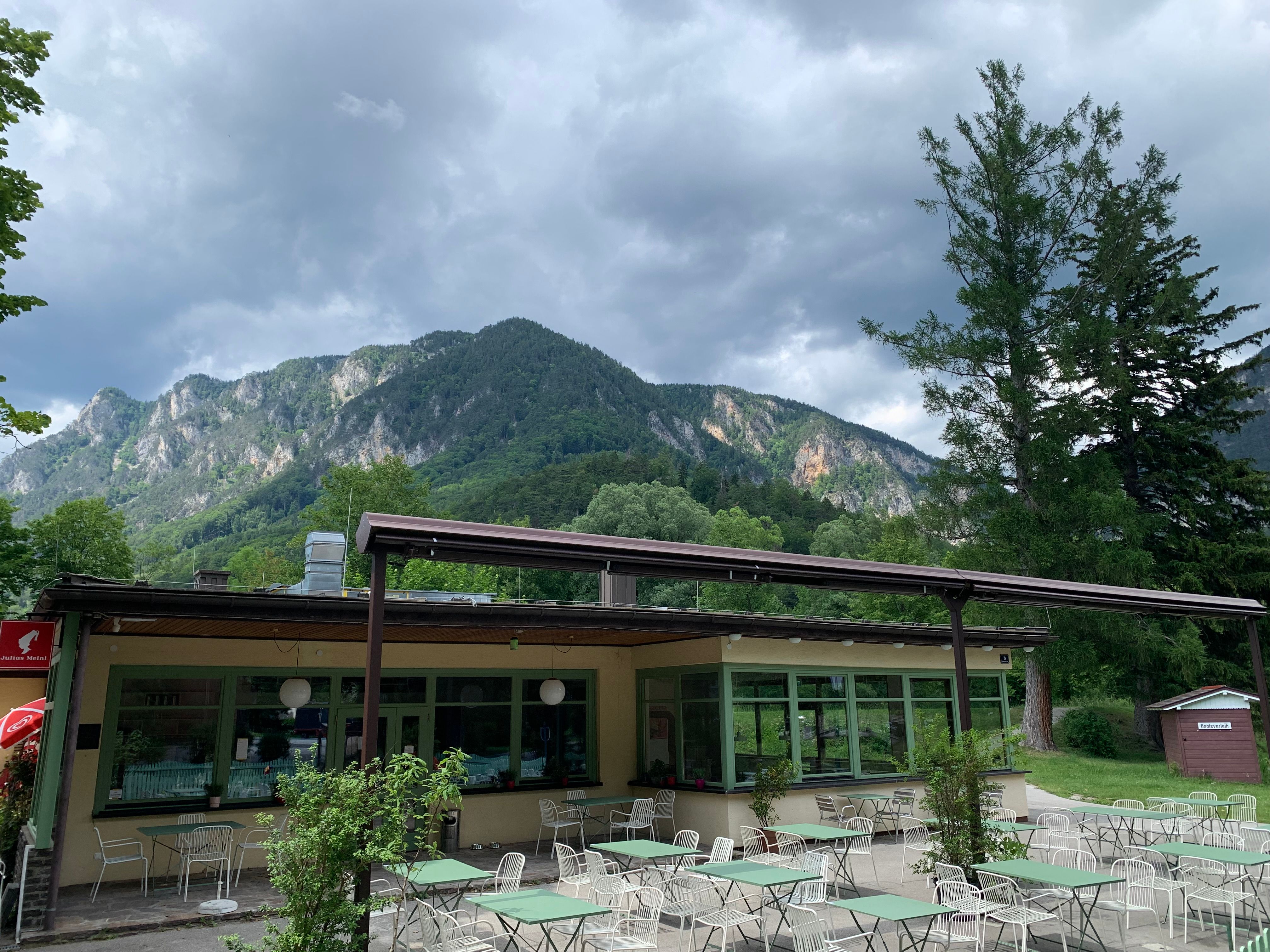 Ein Café mit Terrasse und Tischen im Freien, umgeben von grünen Bäumen und Bergen im Hintergrund.