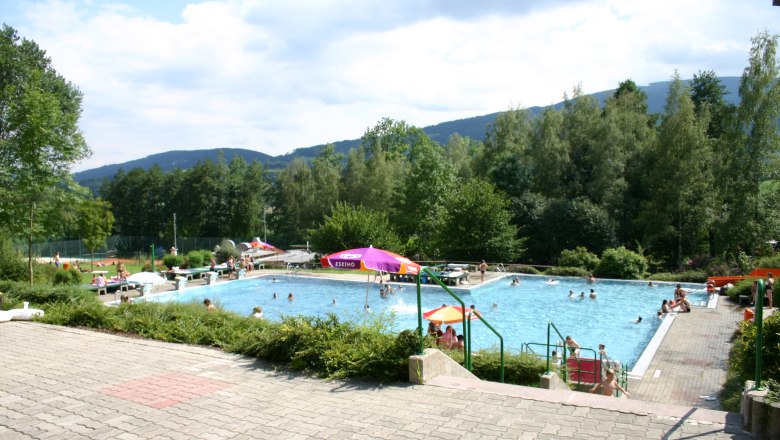 Freibad mit Schwimmern und Sonnenschirmen, umgeben von Bäumen und Hügeln.