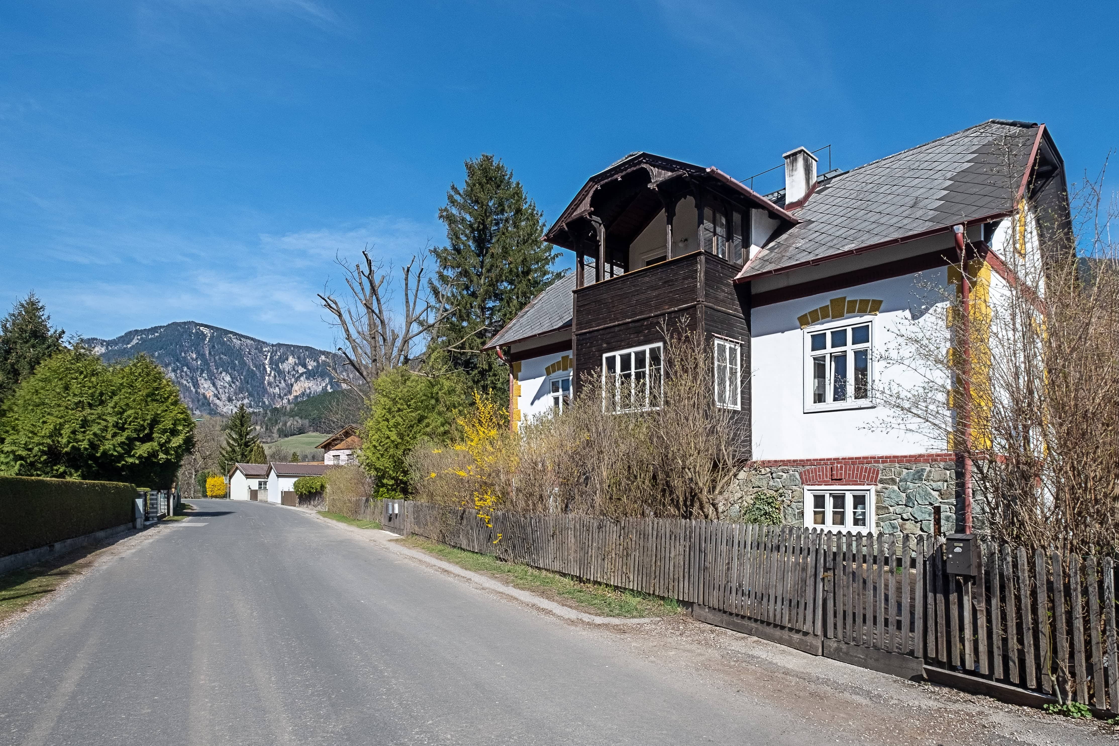 Ein Landhaus mit Holzbalkon und weißer Fassade an einer ruhigen Straße mit Bergblick im Hintergrund.