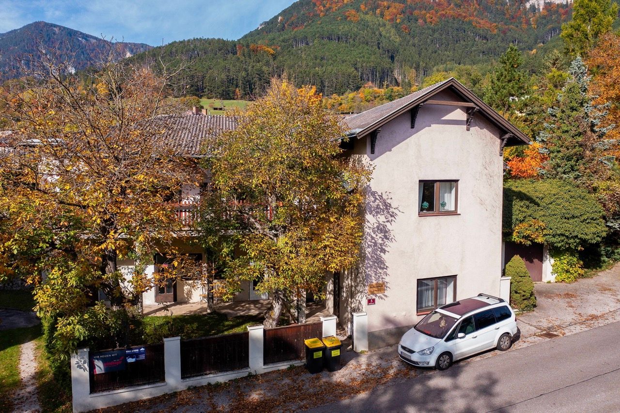 Ein zweistöckiges Gebäude mit Herbstbäumen und einem geparkten Auto davor, umgeben von bewaldeten Hügeln.
