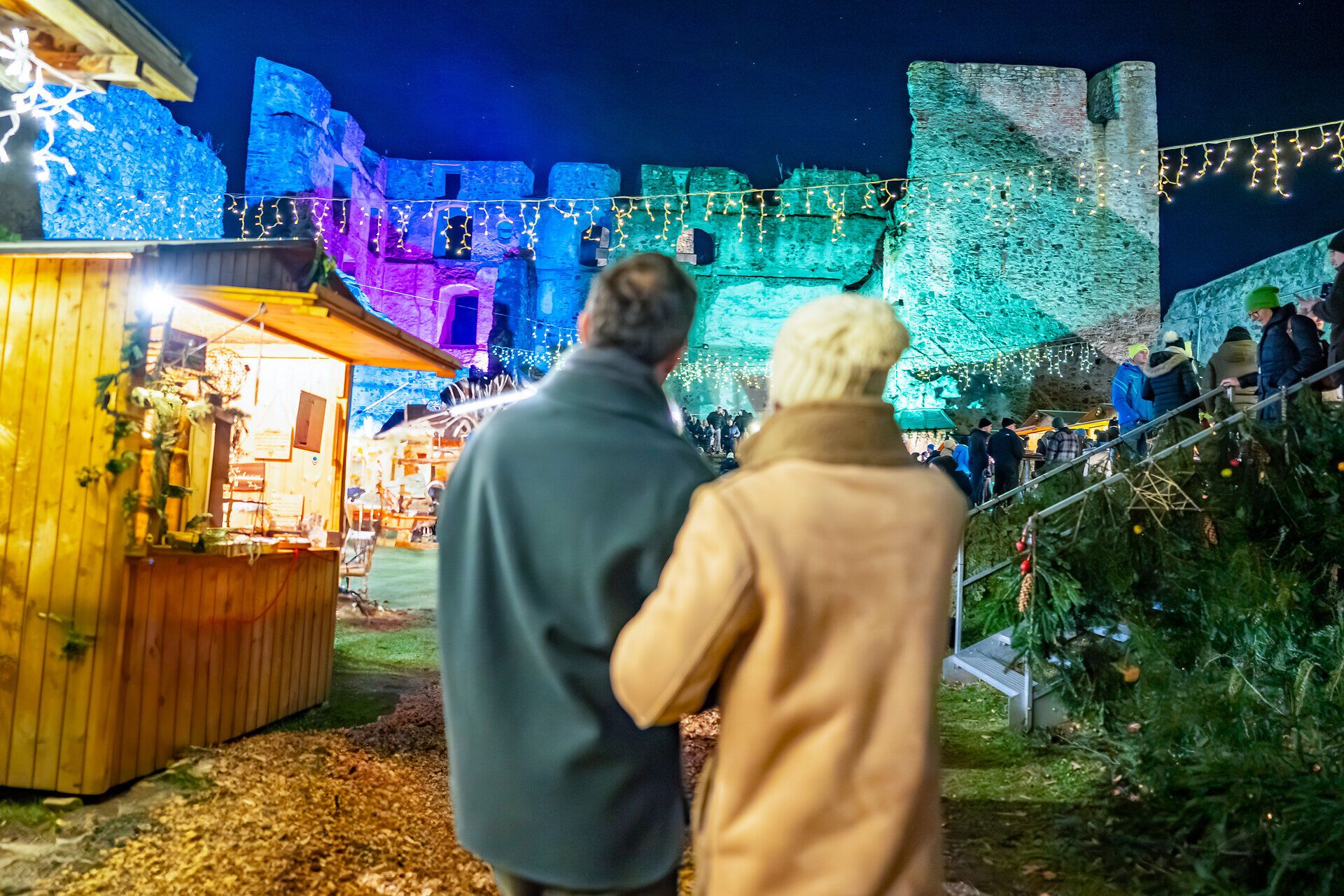 Paar schlendert über einen stimmungsvoll beleuchteten Weihnachtsmarkt vor historischer Burgkulisse bei Nacht.
