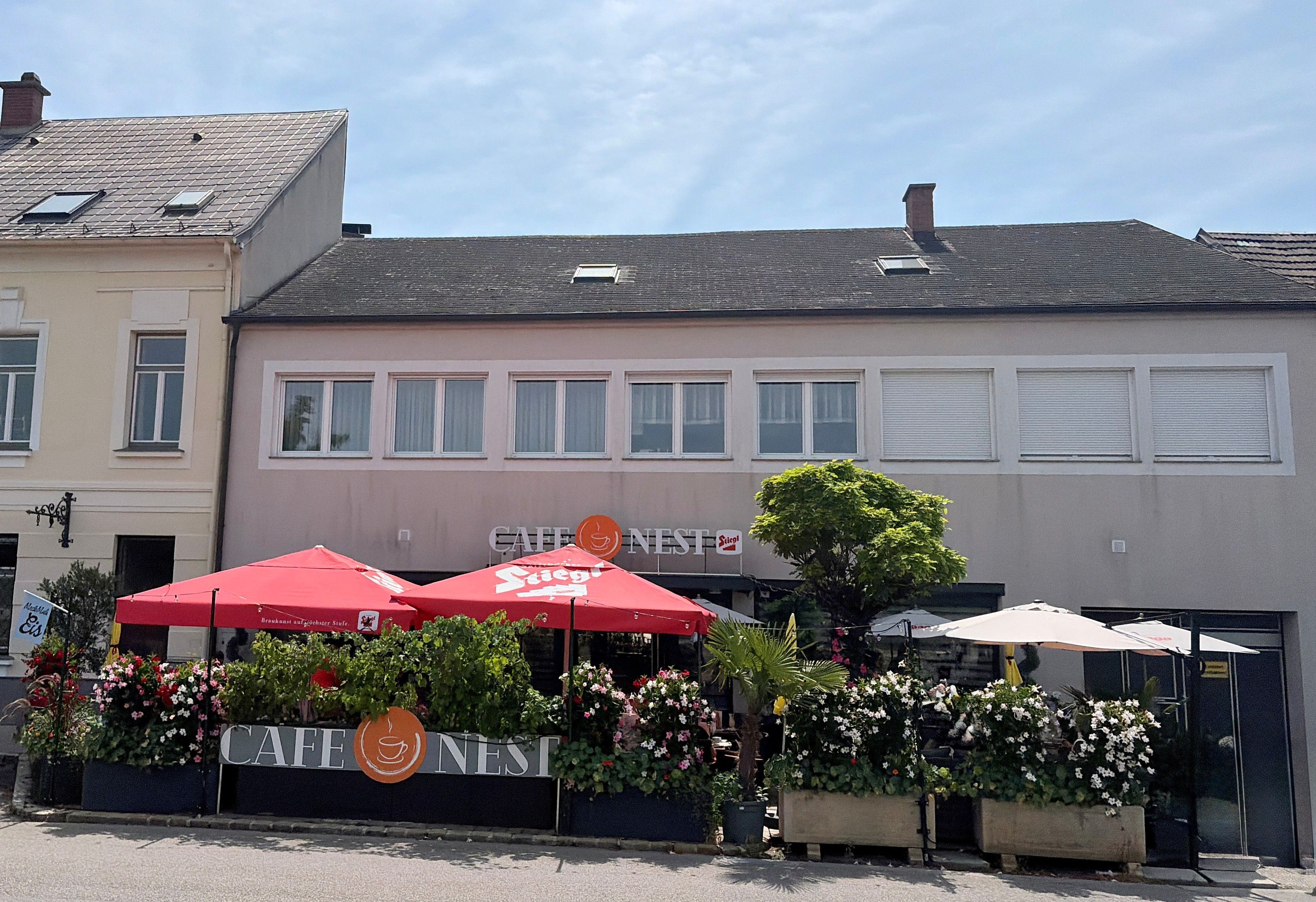 Außenansicht des Cafe Nest in Ternitz mit roten Sonnenschirmen und Pflanzen.