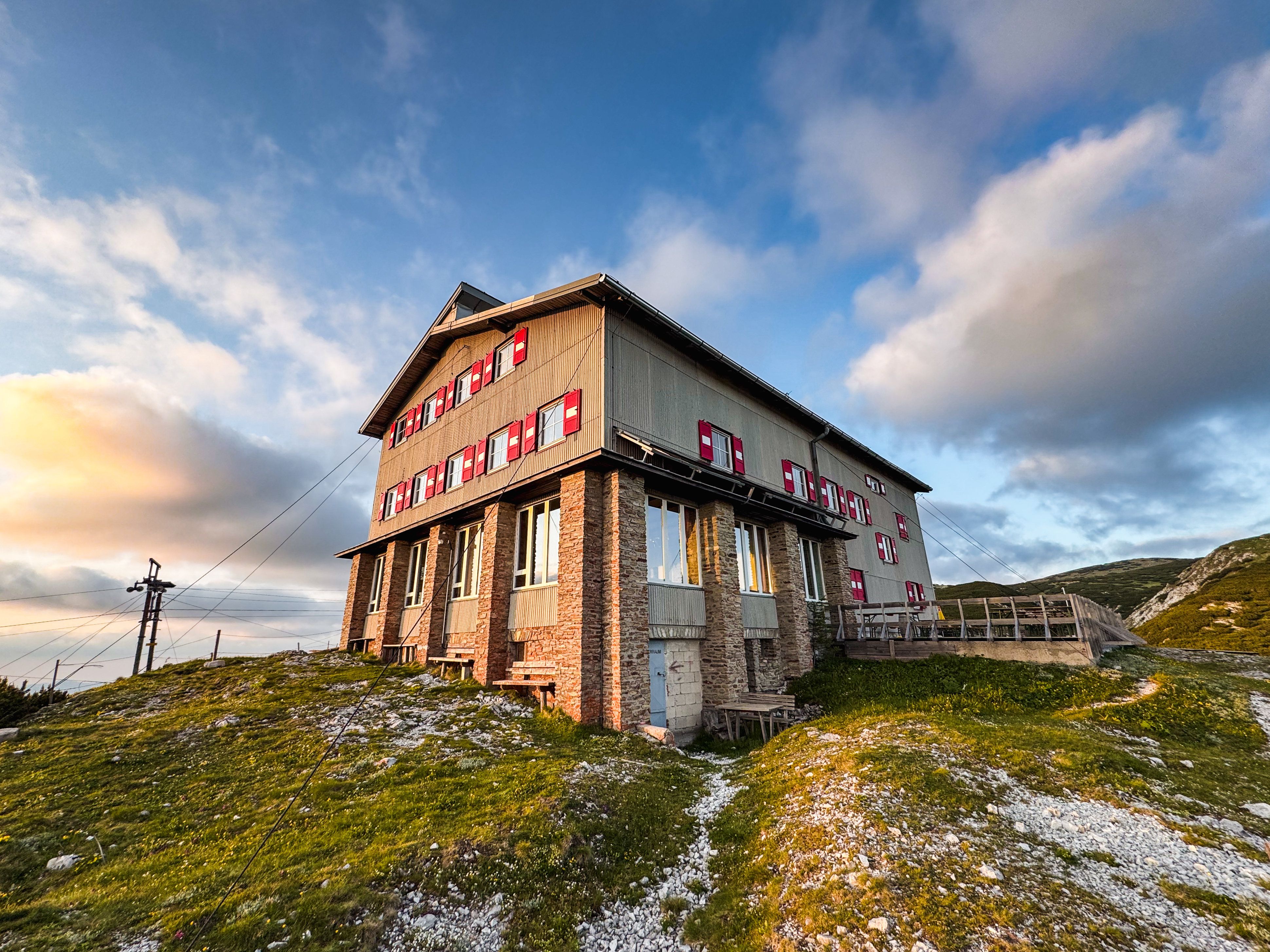 Das Habsburghaus auf der Raxalpe bei Sonnenuntergang, umgeben von grünen Wiesen und einem klaren Himmel mit wenigen Wolken.