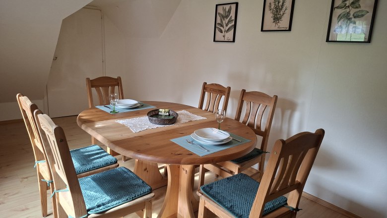 Essbereich mit Holztisch und sechs Stühlen, dekoriert mit blauen Sitzkissen. An der Wand hängen drei botanische Bilder.
