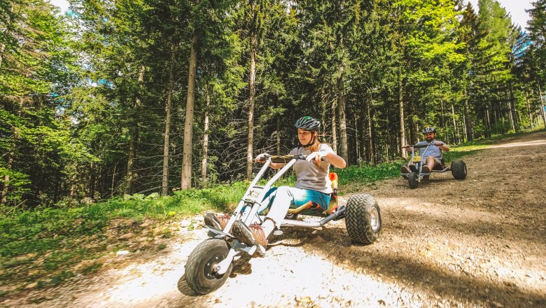 Zwei Personen fahren mit Mountaincarts auf einem Waldweg.