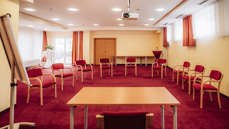 Ein Seminarraum mit rotem Teppich, Stuhlkreis, Tisch, Flipchart und Beamer.