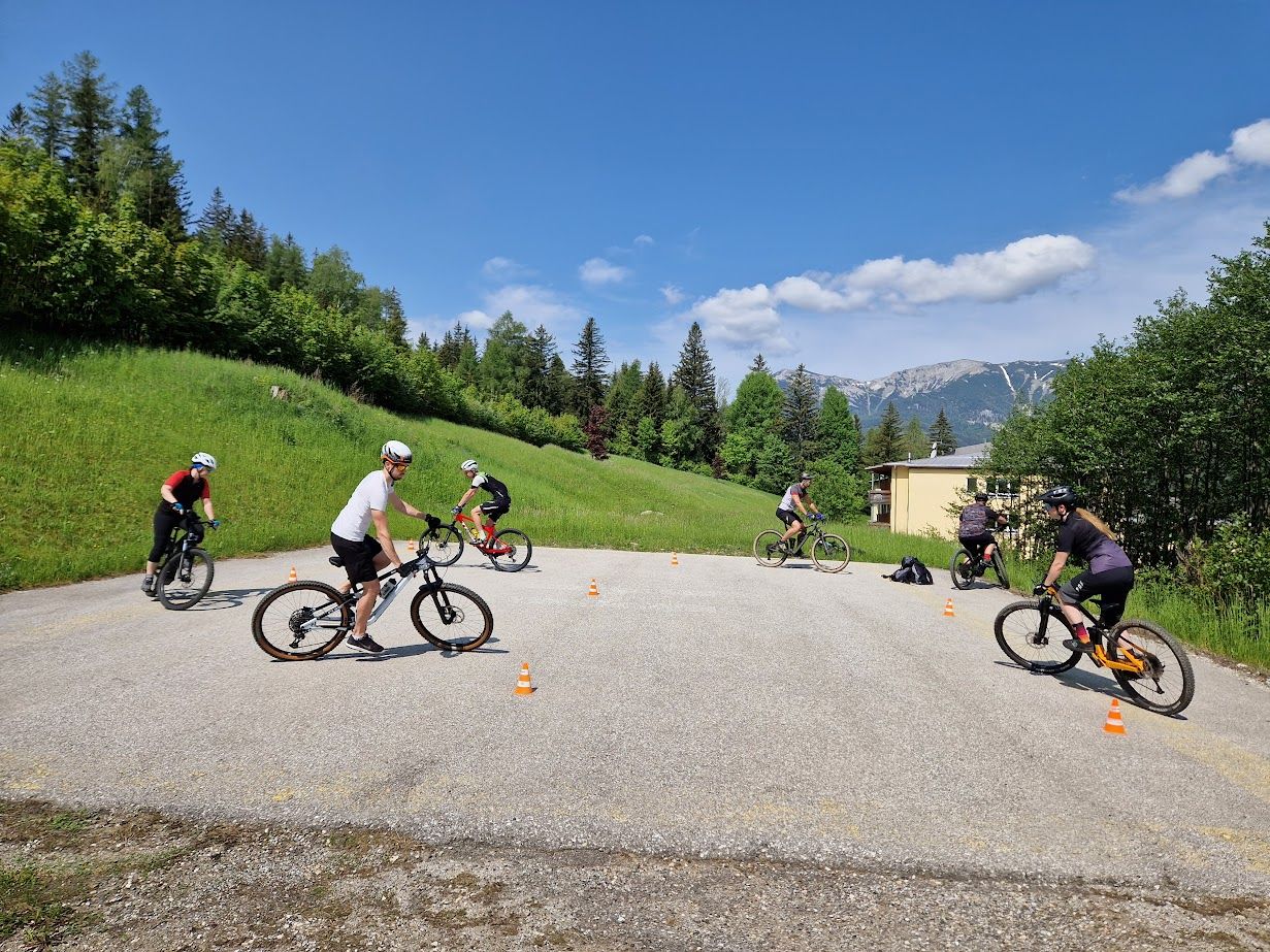 Gruppe von Radfahrern übt auf einem asphaltierten Platz mit Kegeln, umgeben von grünen Hügeln und Bäumen, im Hintergrund Berge.