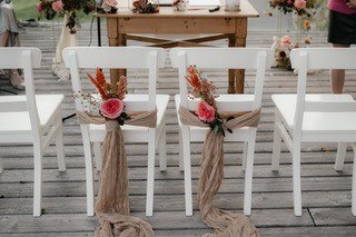 Weiße Stühle mit Blumen und Stoff dekoriert auf einer Holzterrasse, vorbereitet für eine Hochzeit.