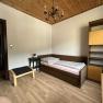 A simply furnished bedroom with single bed, wooden ceiling, shelf and small table with lamp.