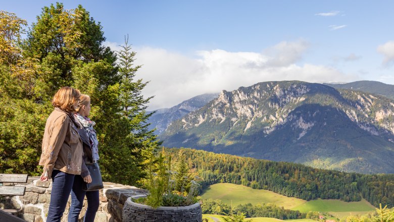 Zwei Personen stehen auf einer Terrasse und blicken auf eine Berglandschaft mit Wäldern und Wiesen.