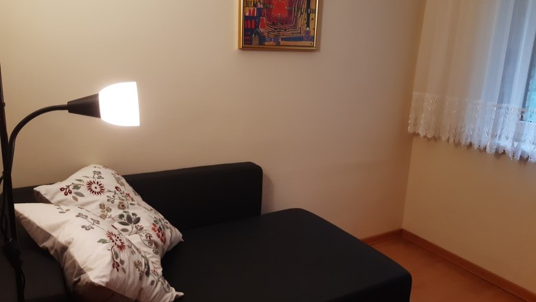 Gem&uuml;tliche Leseecke mit schwarzem Sofa, Stehlampe und dekorativem Kissen. An der Wand h&auml;ngt ein buntes Bild, daneben ein Fenster mit Gardine.