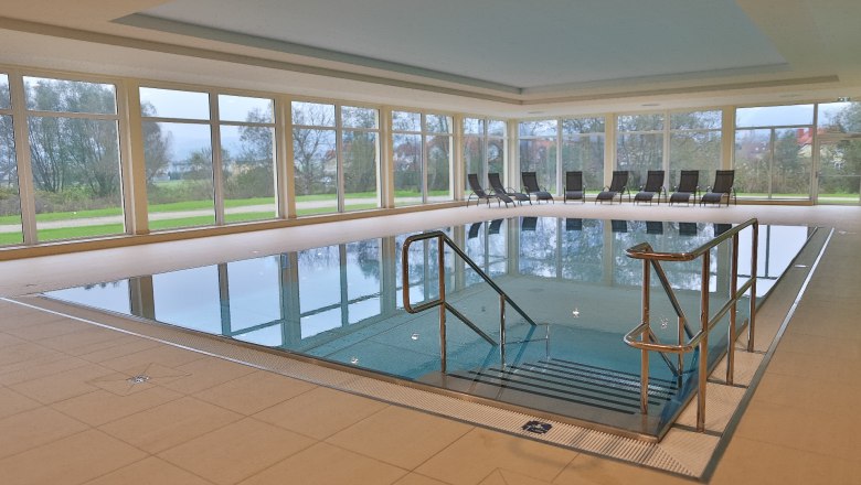 Innenansicht eines Hallenbads mit gro&szlig;en Fenstern und Liegest&uuml;hlen am Beckenrand.