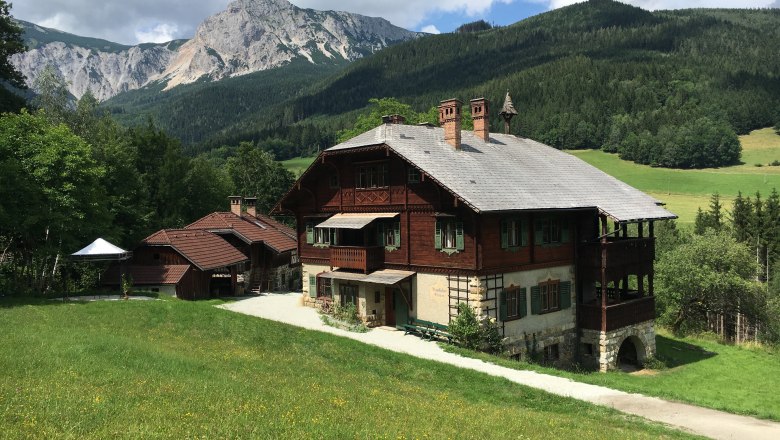 Ein traditionelles Holzhaus in einer bergigen Landschaft mit grünen Wiesen und bewaldeten Hügeln im Hintergrund.
