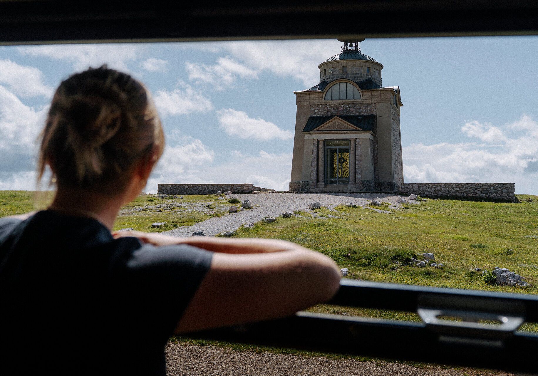 Eine Frau blickt von der Schneebergbahn auf das Elisabethkircherl am Hochschneeberg. Ein Kiesweg führt zur Kirche, die umgeben von einer grünen Wiese ist, was eine Atmosphäre der Erkundung und historischer Neugier erzeugt.