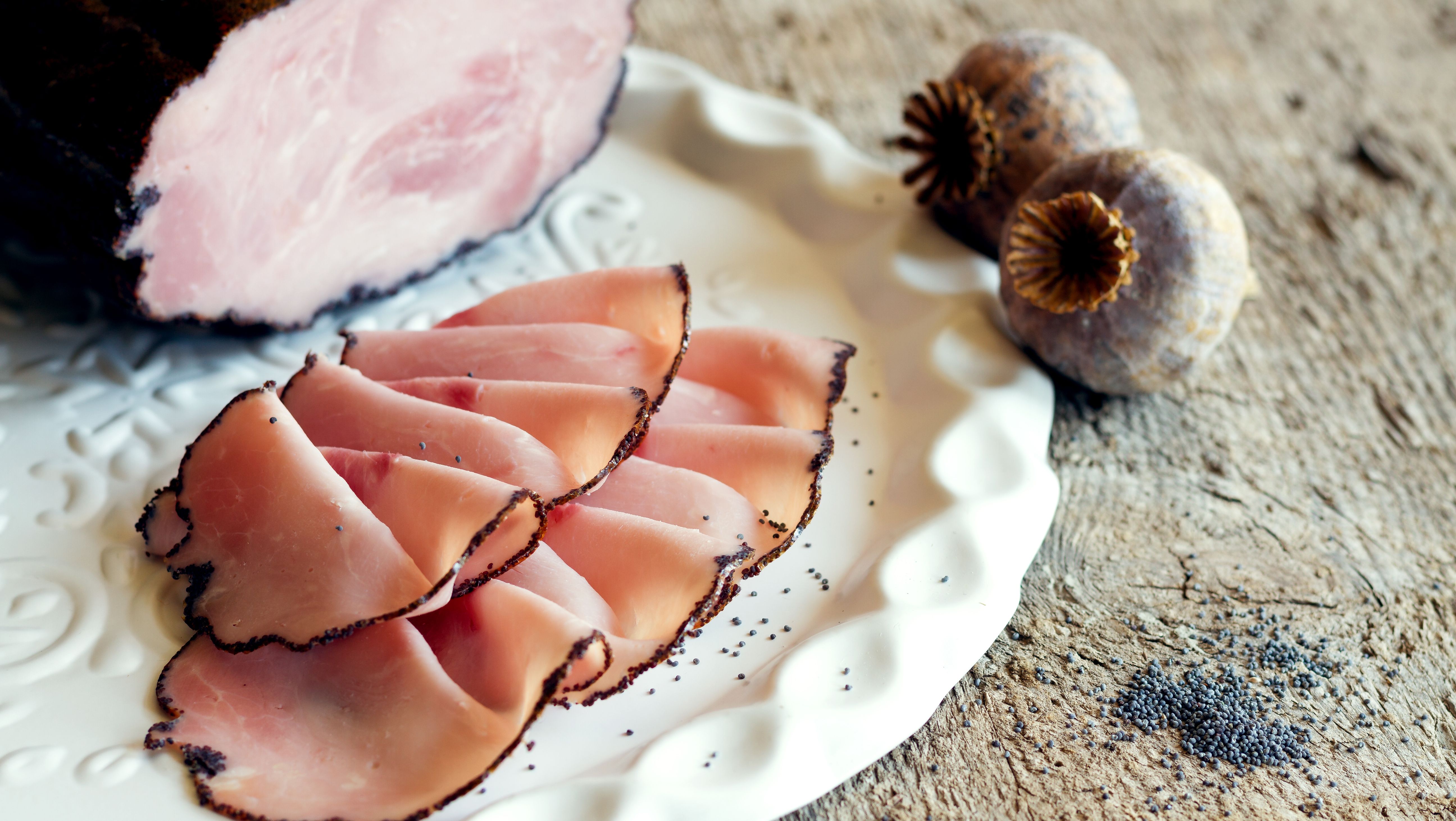 Mohnschinken in Scheiben und im Ganzen auf einem weißen Teller, daneben Mohnkapseln.