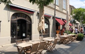 Außenbereich eines Cafés mit Holztischen und Stühlen, roter Markise und Menschen im Hintergrund.