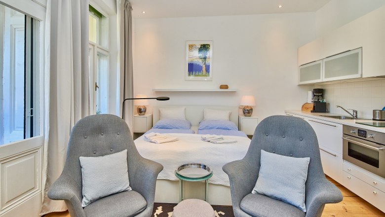 Modernes Studio-Apartment mit Doppelbett, zwei grauen Sesseln, Küchenzeile und großen Fenstern.