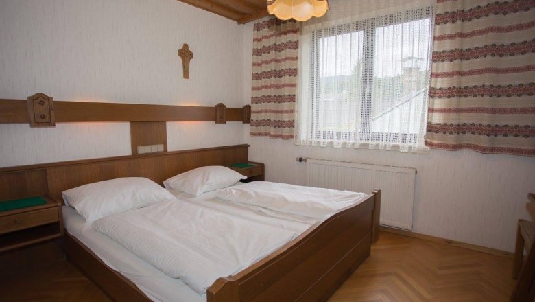 A traditionally furnished double room with wooden furniture, a double bed, a cross on the wall and a window with curtains.