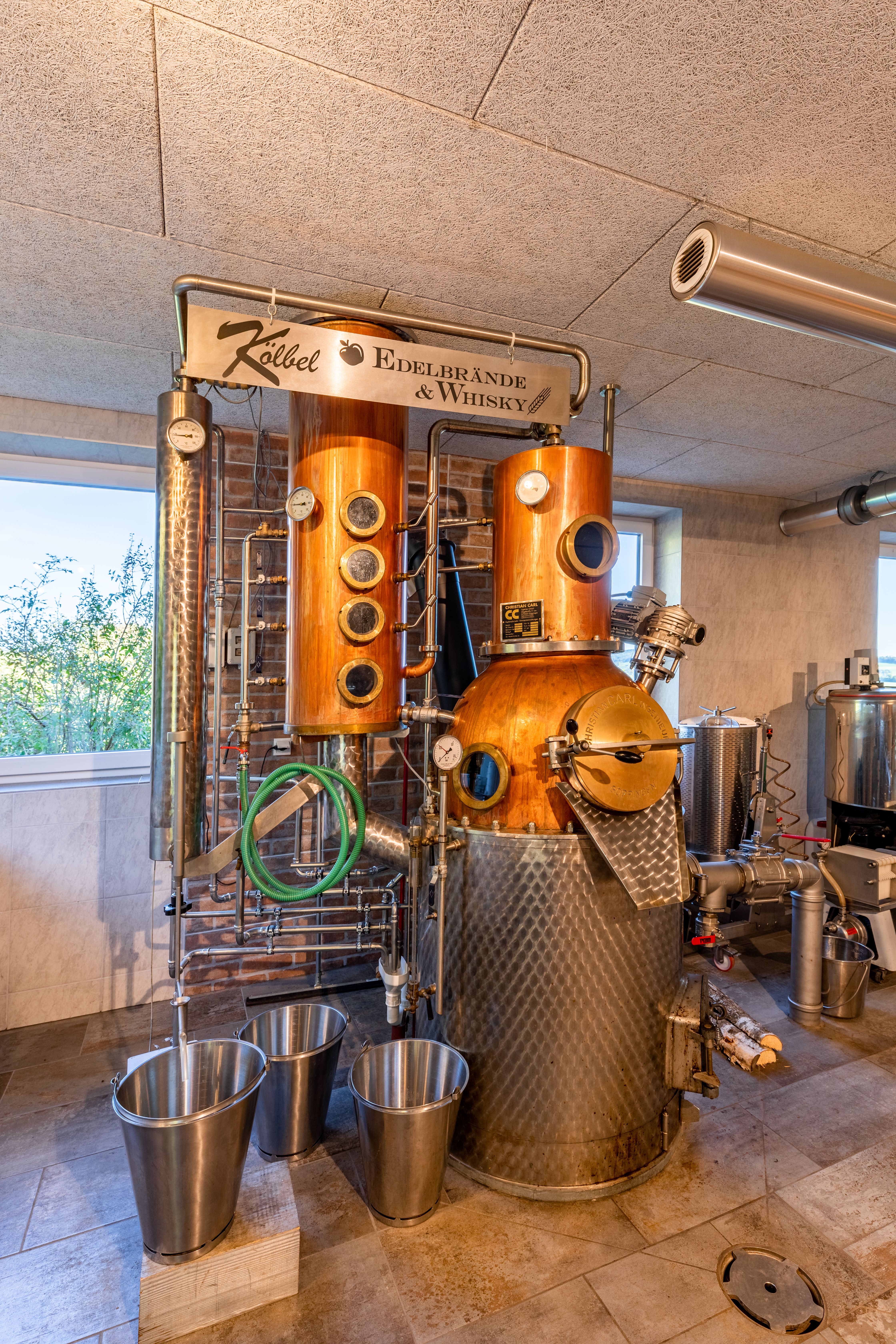 Eine aus Edelstahl und Kupfer gefertigte Destilliermaschine in einem Raum mit Fliesenboden. Viele Regler, Rohre und kleine Schaufenster ins Innere befinden sich an der Maschine. Man sieht auch große Kübel, Holzscheite und das Logo von Kölbel Edelbrände und Whisky auf einem Schild.