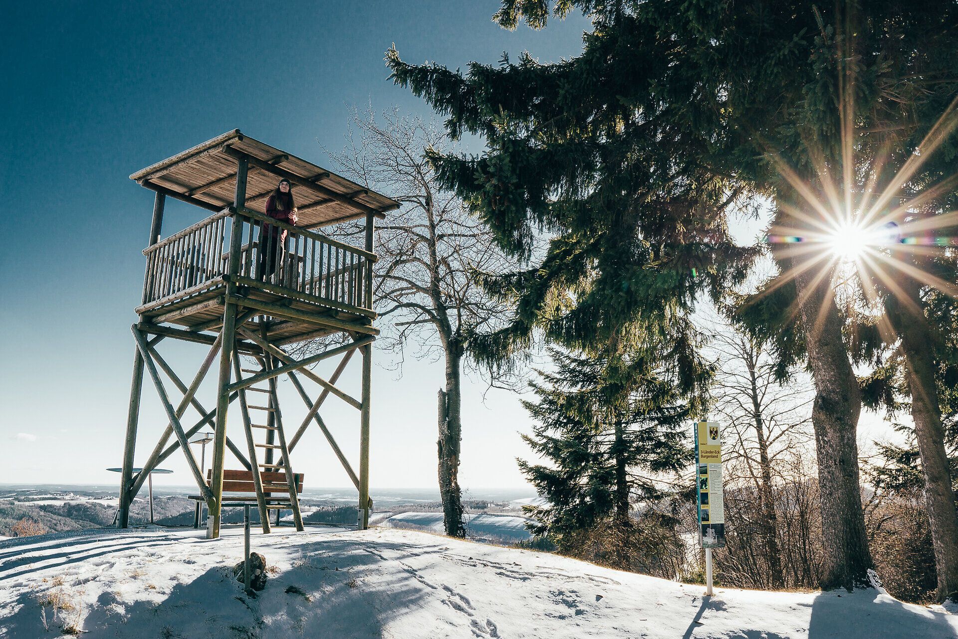 Die winterliche Landschaft strahlt in einem glitzernden Weiß, während die Sonne sanft hinter den Bäumen aufgeht. Von der Aussichtswarte aus eröffnet sich ein atemberaubender Blick über die verschneiten Hügel und Wälder, die zum Verweilen und Entdecken einladen.