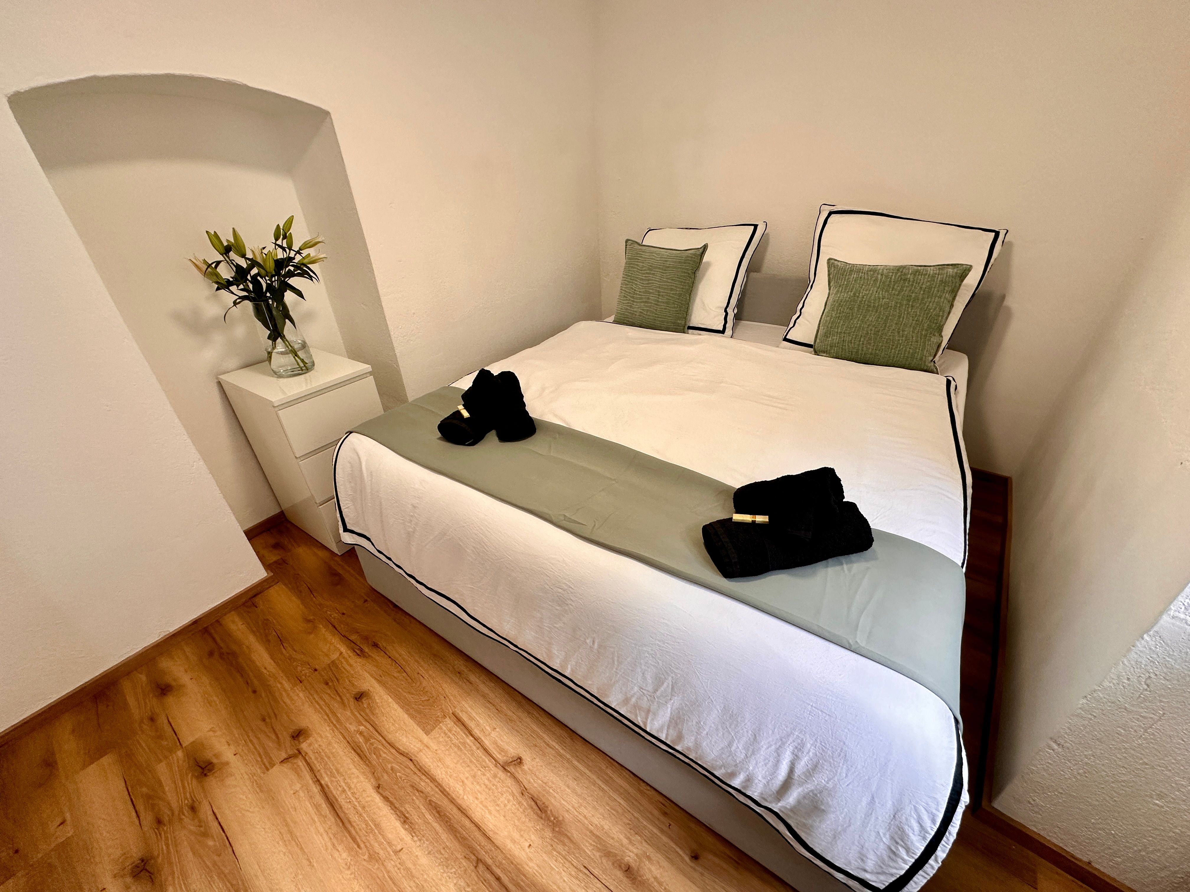 Ein gemütliches Schlafzimmer mit einem Doppelbett, weißen und grünen Kissen, einem Nachttisch mit Blumen und einem Holzboden.