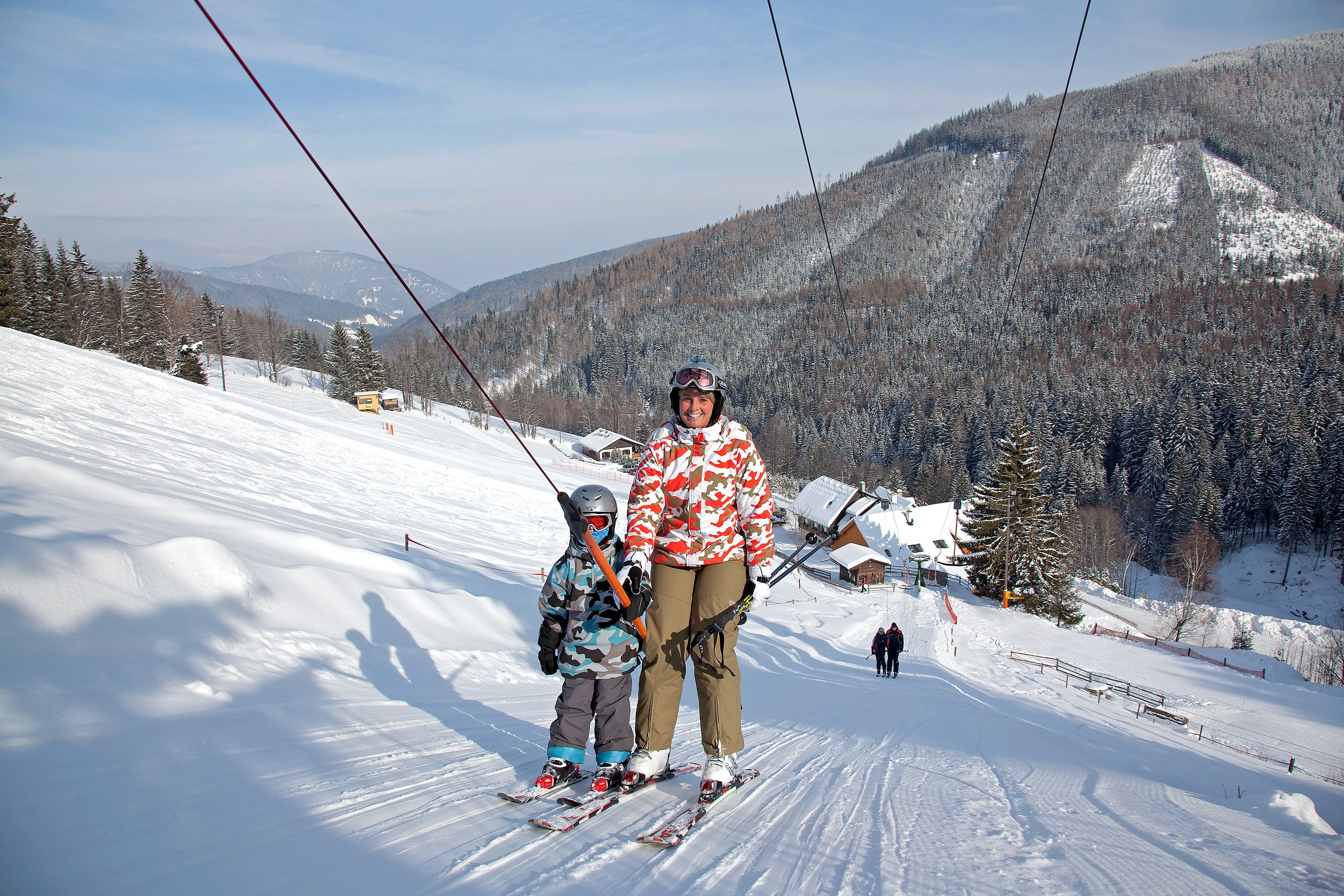 Zwei Skifahrer auf einem Schlepplift in einer verschneiten Berglandschaft.