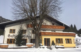 Ein Gasthof im Schnee mit Holzverzierungen und einem großen Baum davor.