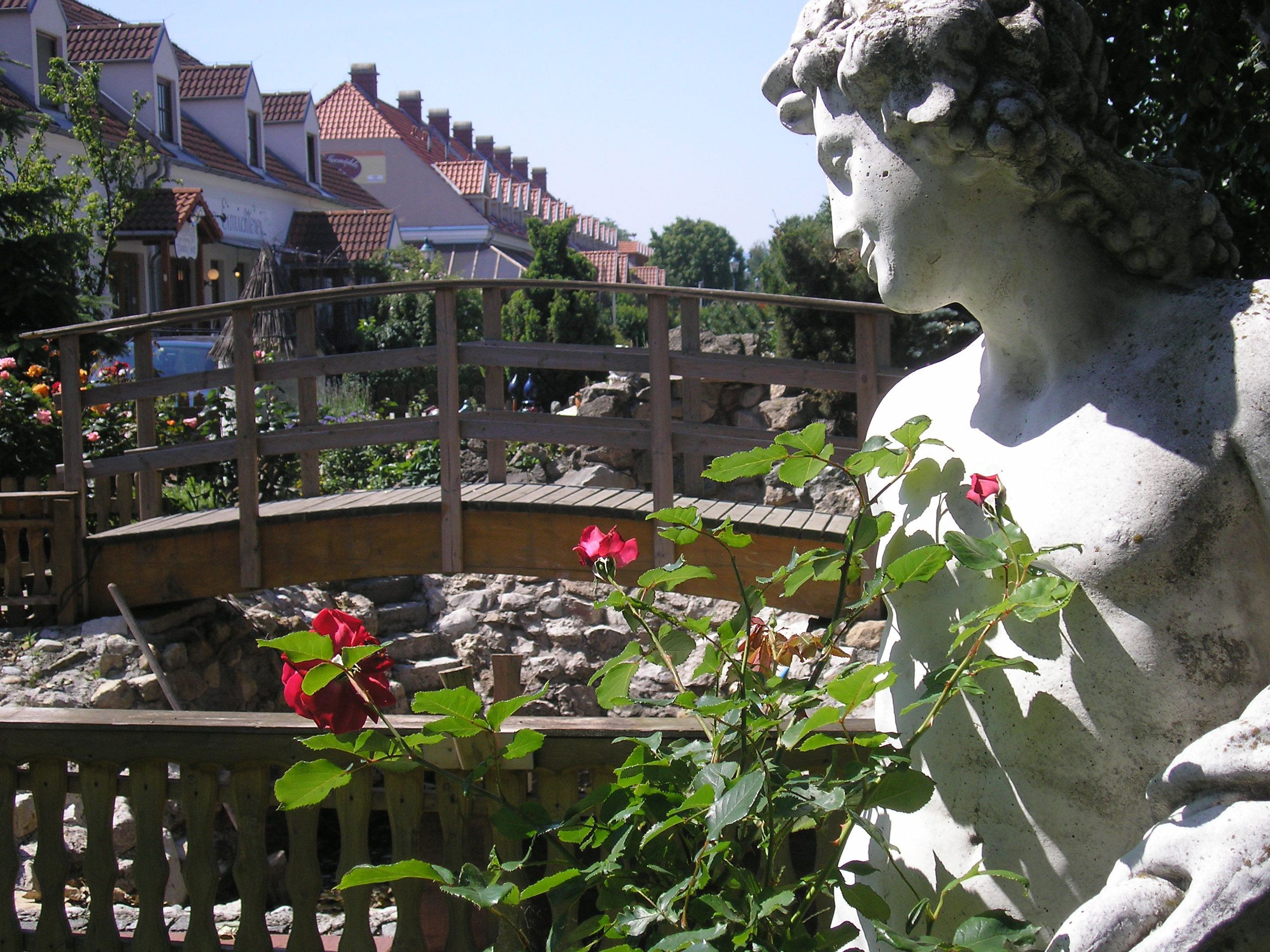 Steinstatue neben einer Holzbrücke und roten Rosen im Vordergrund.