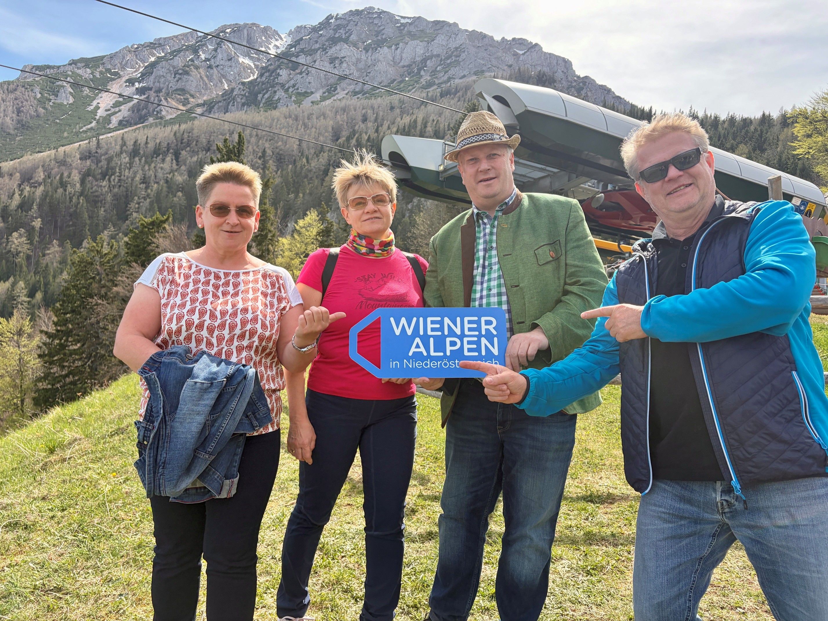 Vier Personen stehen vor einer Sessellift-Bergstation. Eine Person hält ein Wiener Alpen-Logo Schild in den Händen.