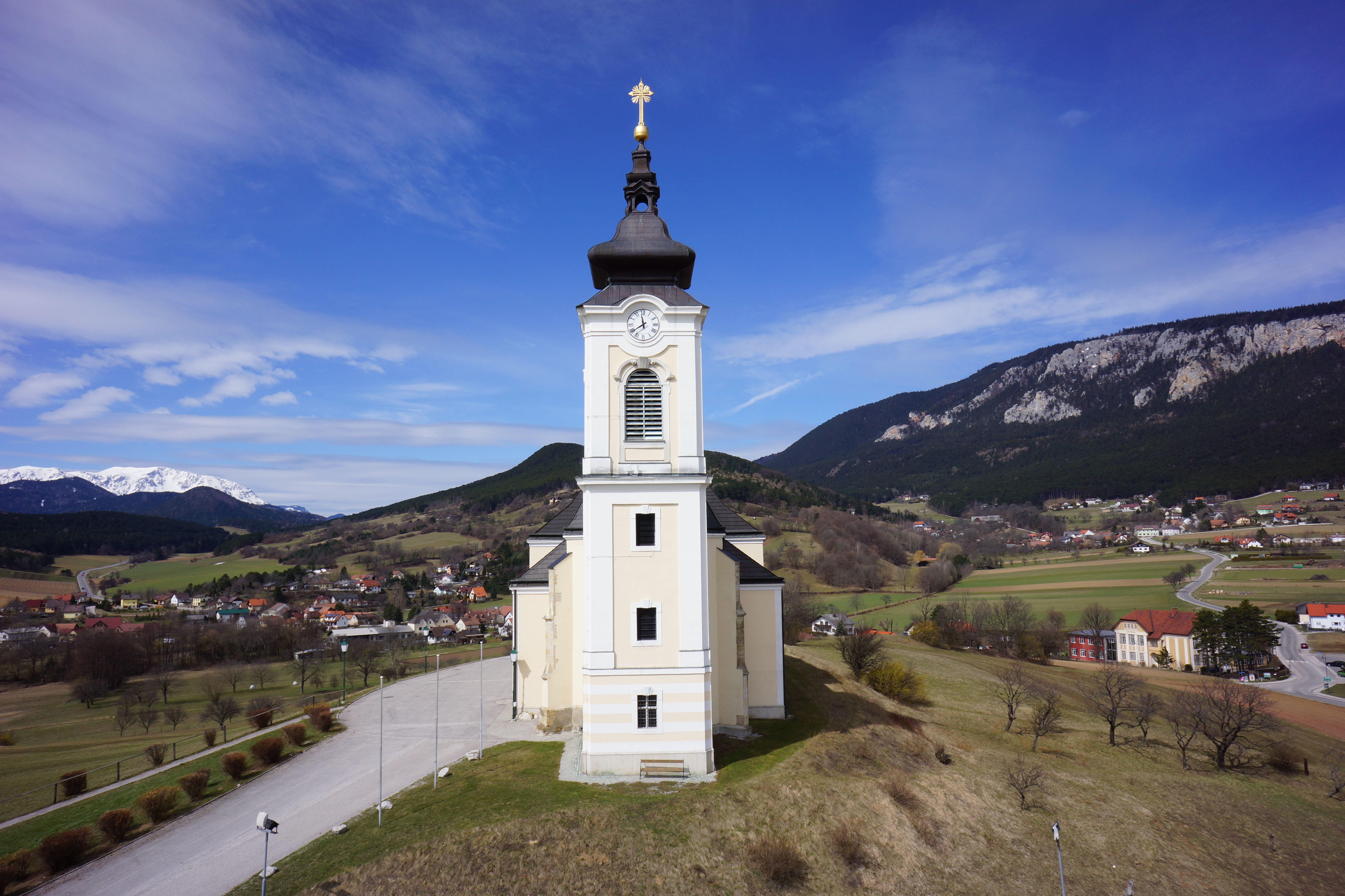 Kirche mit Uhrturm in ländlicher Landschaft mit Bergen im Hintergrund.