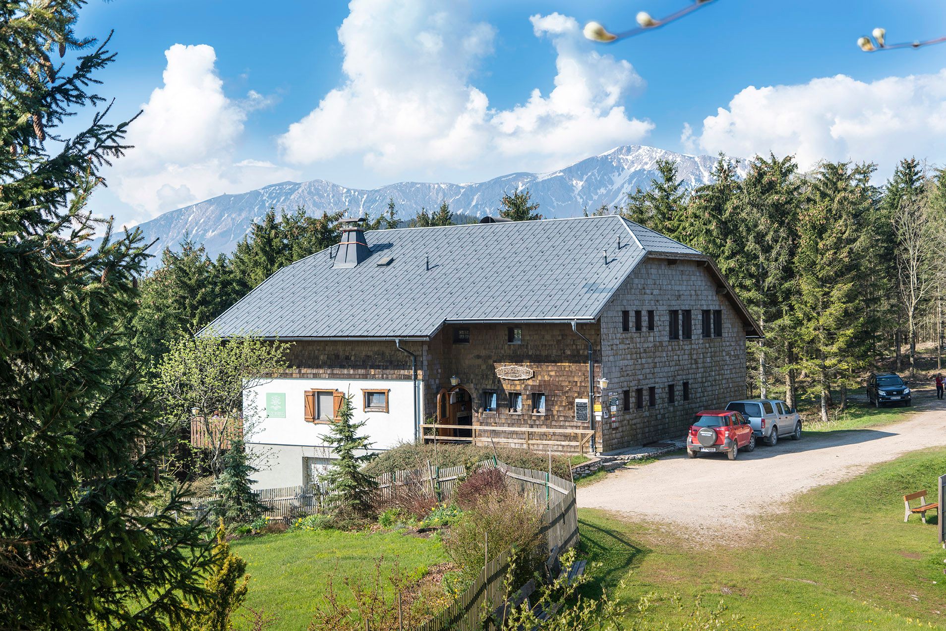 Eine Berghütte mit grauem Dach, umgeben von Bäumen und Bergen im Hintergrund.