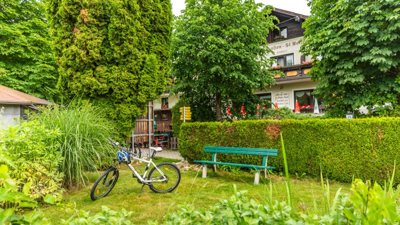 A bicycle is parked next to a bench in the garden of an inn, surrounded by trees and bushes.
