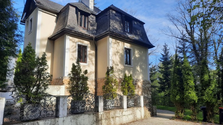 Eine zweistöckige Villa mit Mansardendach, umgeben von Bäumen und einem schmiedeeisernen Zaun.