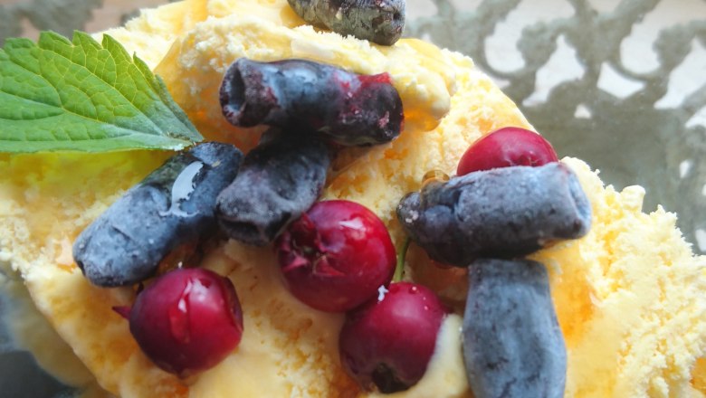 Ein Dessert mit gelbem Eis, roten und blauen Beeren, garniert mit einem grünen Blatt, in einer Glasschale.