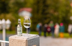 Bottle and glass with Silva Verjus drink on a concrete pillar, blurred people in the background.