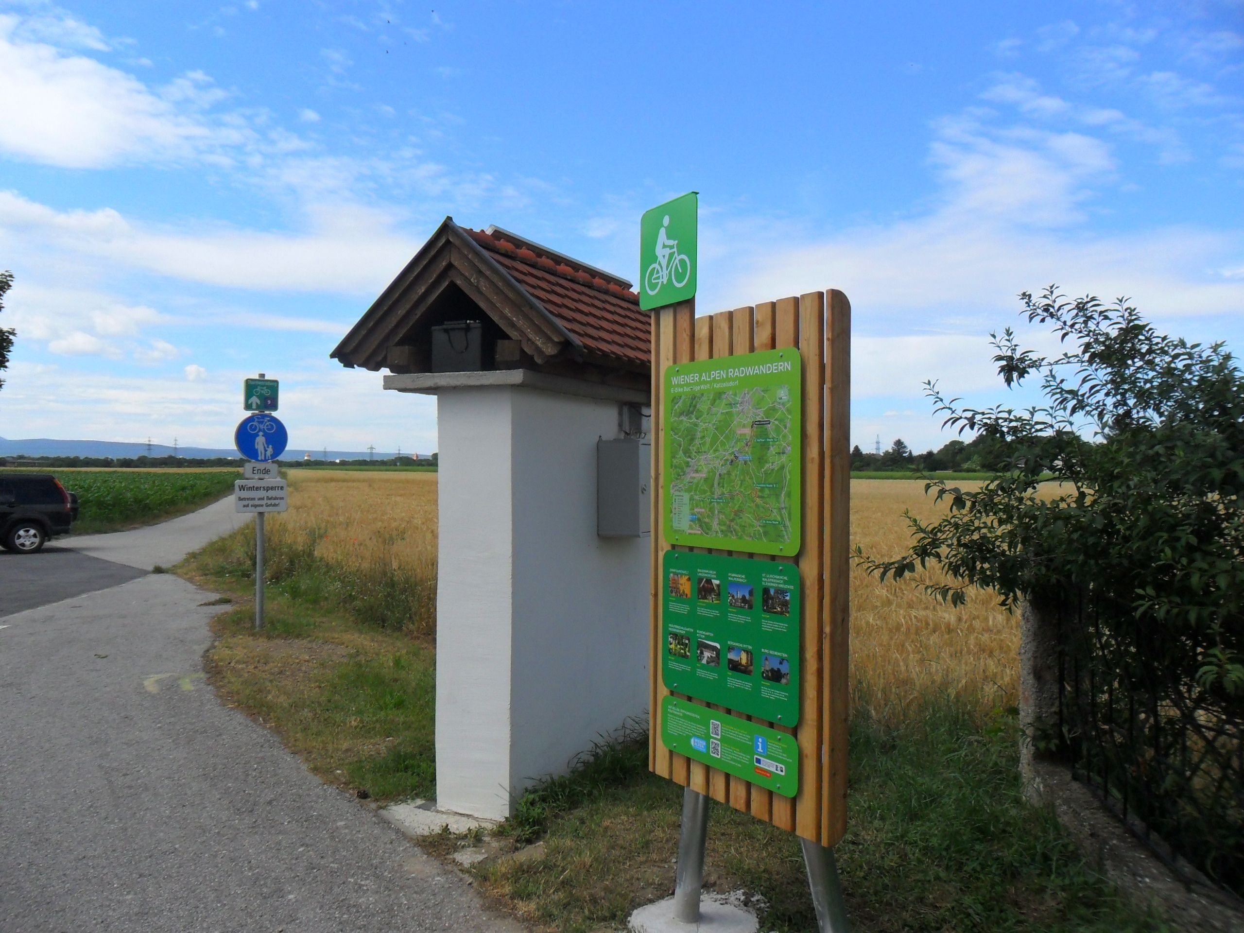 Radstartplatz-Tafel in Katzelsdorf mit Wegweiser und Landschaft im Hintergrund.