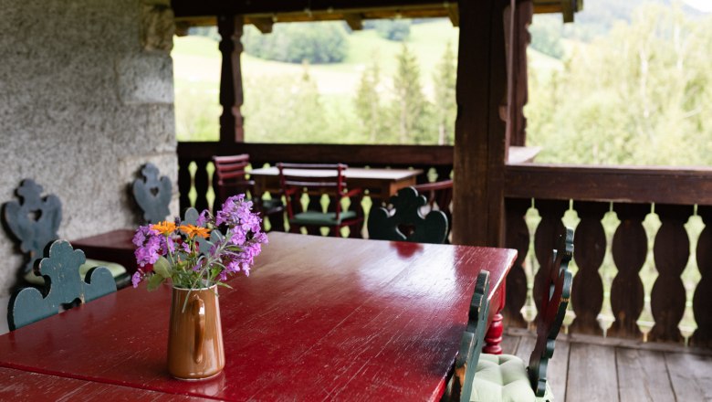 A rustic veranda with wooden tables and chairs, decorated with a jug full of colorful flowers. A green landscape can be seen in the background.
