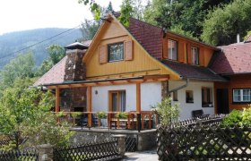Ein traditionelles Ferienhaus mit Holzveranda und Schindeldach, umgeben von Bäumen und Bergen.