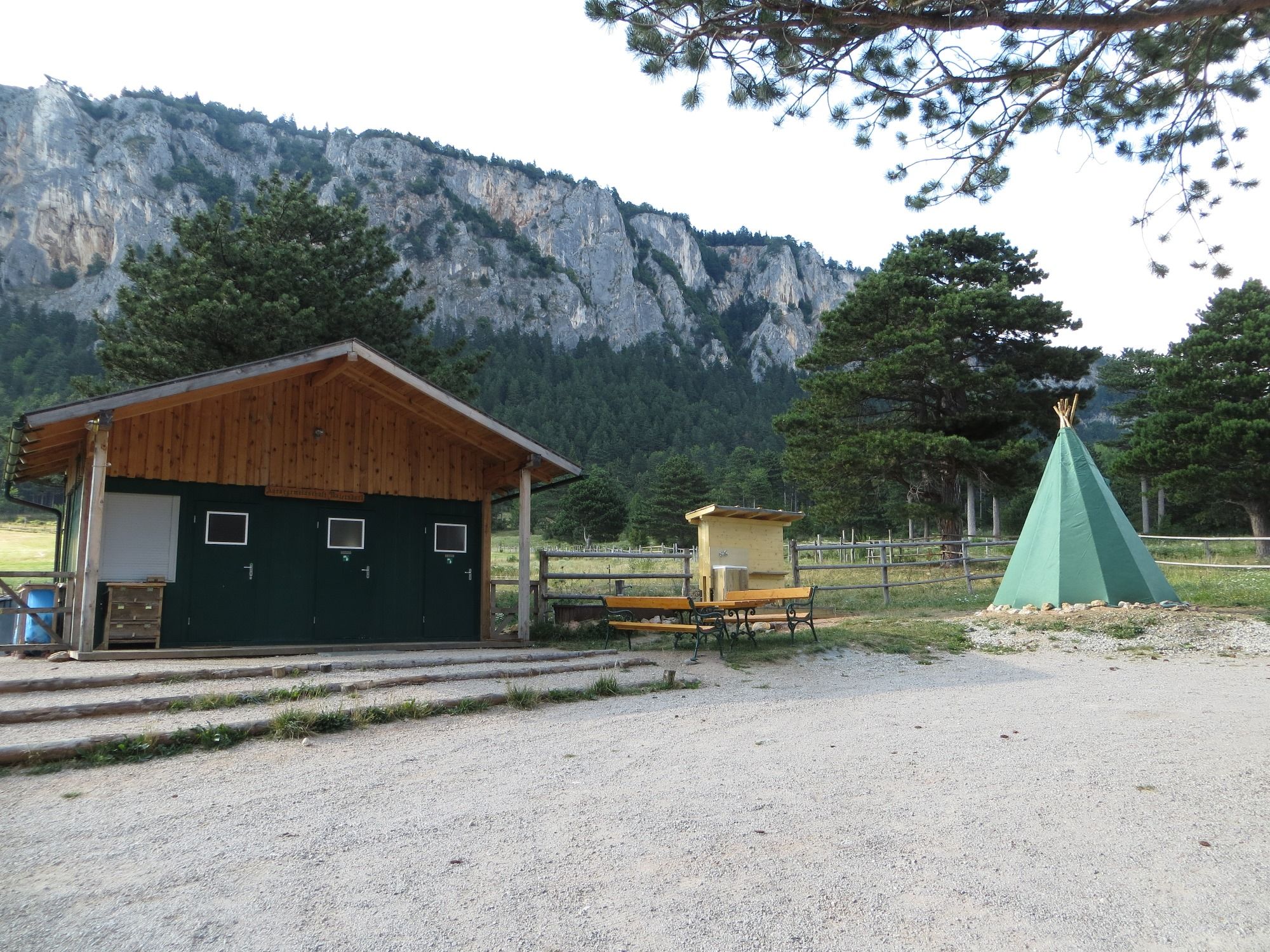 Ein Zeltplatz mit einem grünen Tipi, einer Holzhütte und Bänken vor einer Felswand und Bäumen.