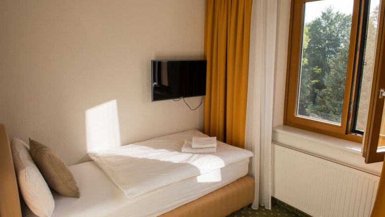 Einzelzimmer mit Einzelbett, gelben Vorhängen, einem Fenster mit Blick ins Grüne und einem an der Wand montierten Fernseher.