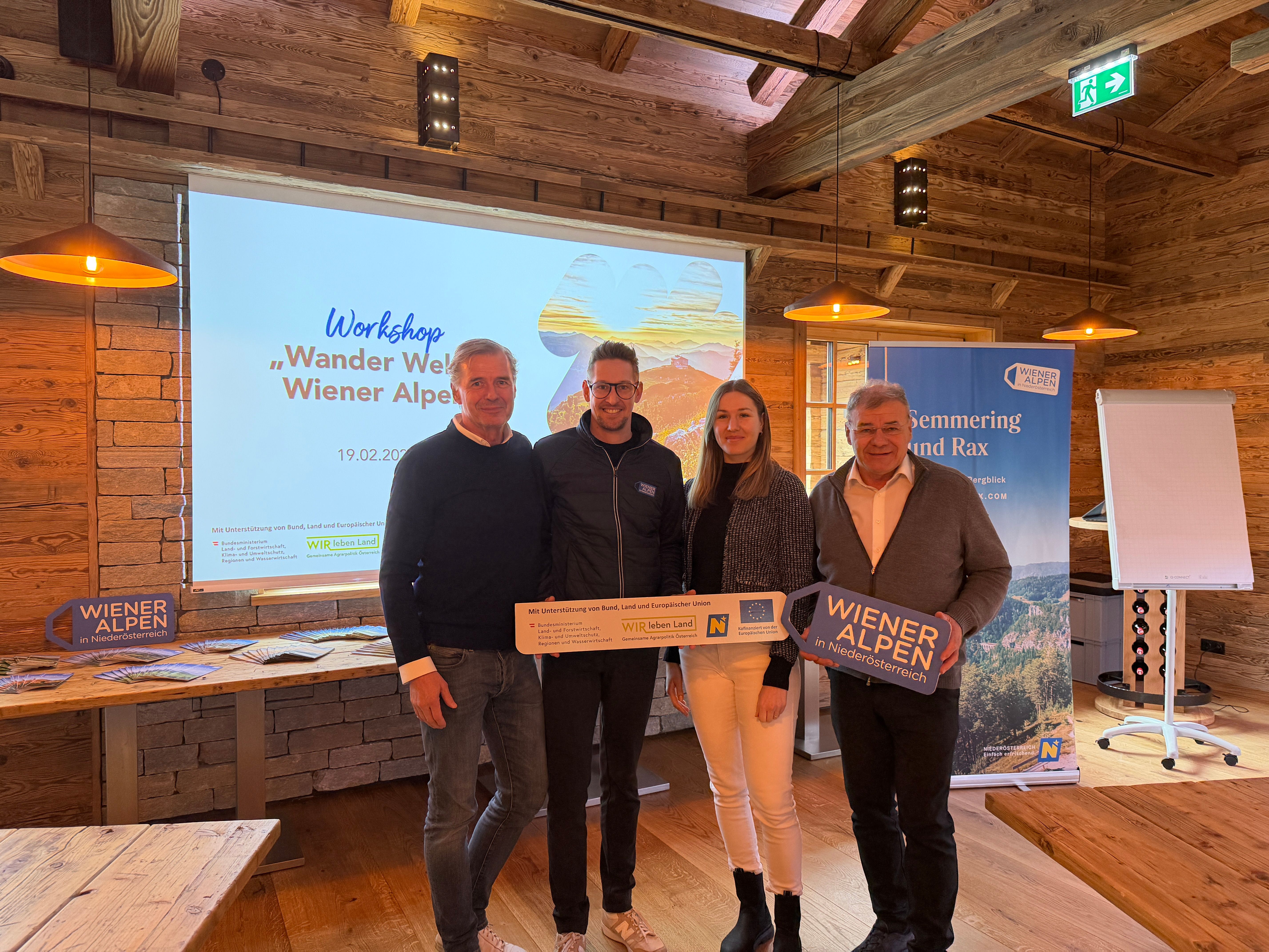 Vier Personen stehen in einem rustikalen Raum vor einer Leinwand nebeneinander und halten ein Wiener Alpen Schild sowie ein LEADER-Schild in den Händen. 