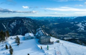 Aerial view of the Raxalm mountain inn in winter with snow-covered mountains and valleys in the background.