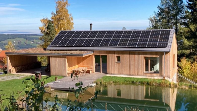 Ein modernes Holzhaus mit Solarpanelen auf dem Dach, umgeben von Natur und einem kleinen Teich im Vordergrund.