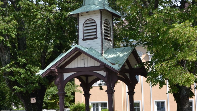 Ein kleiner Pavillon mit spitzem Dach und Kreuz, umgeben von Bäumen.