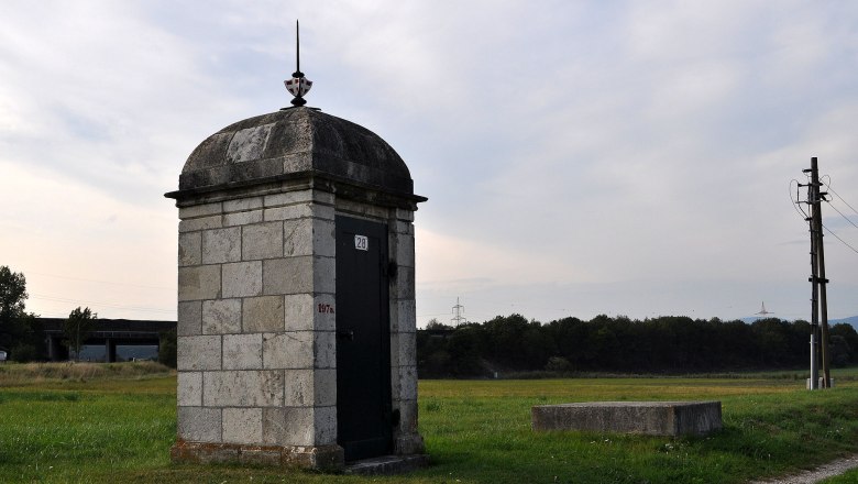 Steinbauwerk auf einer Wiese mit Strommast im Hintergrund.