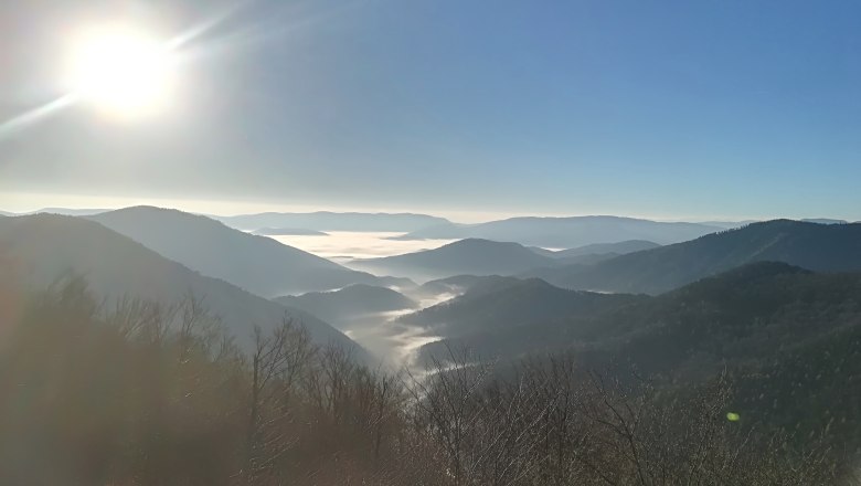 Blick von einem Berg auf nebelverhangene Täler und Hügel unter strahlend blauem Himmel mit Sonnenschein.